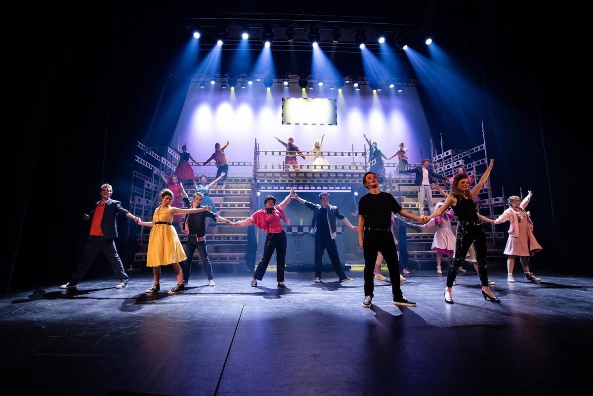 Elever fra Karmøy kulturskole opptrer på en opplyst scene under en musikalforestilling i Festiviteten. Flere dansere og skuespillere står spredt på scenen med armene løftet, mens scenelys lyser ned fra taket. - Klikk for stort bilde